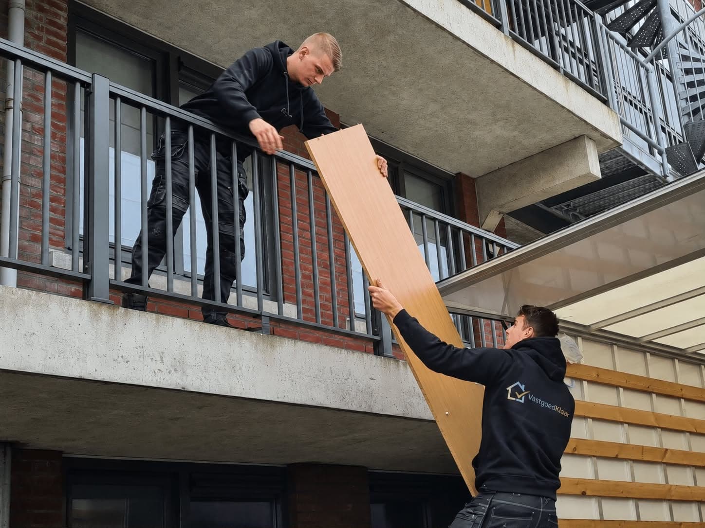 Twee medewerkers van VastgoedKlaar dragen een houten paneel omhoog bij een appartement